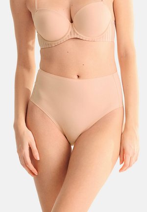 Femme portant une lingerie beige taille haute et un soutien-gorge de maintien assorti, debout devant un fond clair uni.