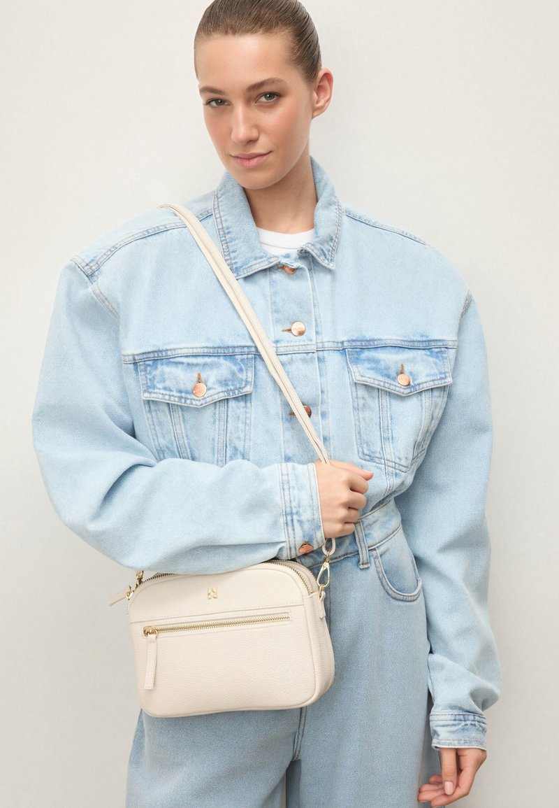 Joven con chaqueta vaquera clara de gran tamaño y vaqueros, sosteniendo un pequeño bolso bandolera crema con cremallera y correa doradas.