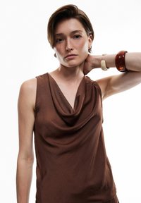 Top sin mangas de color marrón con cuello de pico, hecho de un tejido ligero y sedoso. La modelo lleva una pulsera texturizada en una muñeca. Diseño simple y elegante.