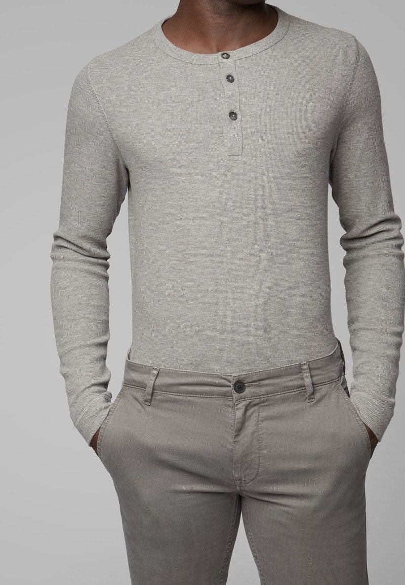 Šedé žebrované tričko s dlouhým rukávem typu henley s třemi knoflíky na krku, v kombinaci s světle šedými kalhotami se zapínáním na předním knoflíku.
