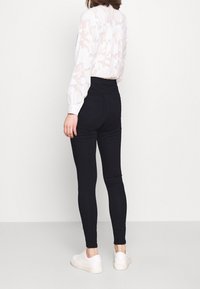 Jeggings negros de talle alto con una textura suave, que cuentan con dos bolsillos traseros, combinados con una blusa translúcida de estampado ligero y zapatillas blancas.