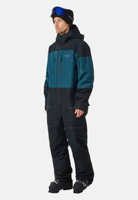 Skistoneel met een donkerblauw en zwart ontwerp, gemaakt van waterdicht materiaal met meerdere ritszakken en een capuchon. Inclusief skibril.