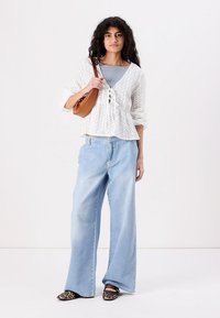 Blusa bianca in pizzo con nodo frontale, canotta a coste azzurro chiaro, jeans a gamba larga in denim chiaro e scarpe slip-on con stampa animale. Borsa marrone.