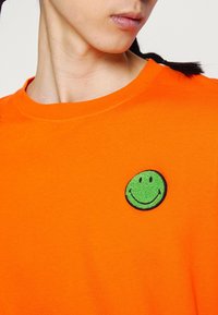 Personne portant une chemise orange avec un petit badge vert en forme de visage souriant sur la poitrine, visible du cou au haut du torse.