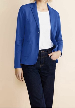 Vrouw draagt een felblauwe blazer over een wit shirt met donkere spijkerbroek, één hand in de zak, tegen een effen beige achtergrond.