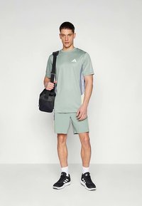 Vaaleanvihreä urheilupaita ja shortsit, joissa on sinisiä ja keltaisia yksityiskohtia. Musta laukku olalla. Mustat urheilukengät.