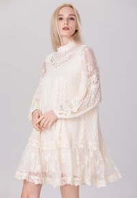 Robe en dentelle de couleur crème avec un col haut, des manches longues transparentes et un ourlet à volants en couches avec des motifs floraux et des détails en dentelle délicate.