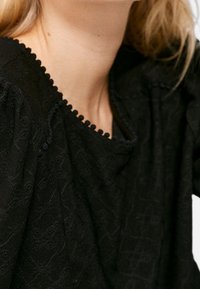 Top negro con tela texturizada, escote con borde ondulado y mangas sueltas. Patrón sutil visible en el material, que añade interés visual.