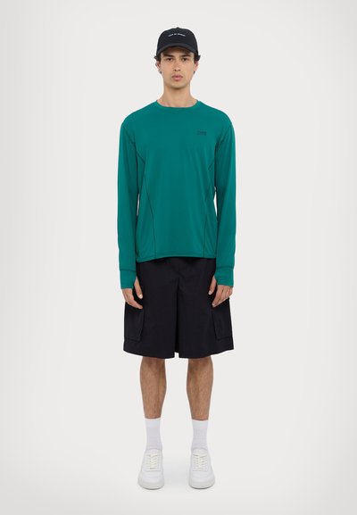 Lange mouw teal shirt met een ronde hals, gecombineerd met zwarte cargoshorts. Witte sneakers en een zwarte baseballcap maken de outfit af.