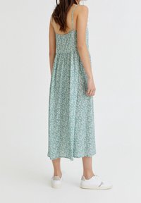 Vestido maxi floral verde con tirantes finos, cintura ceñida y falda fluida. Combinado con zapatillas blancas para un look casual.