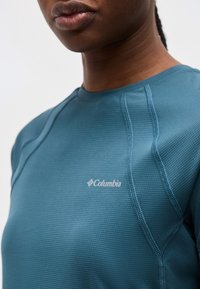 Κοντινό πλάνο ενός ατόμου που φοράει μπλε αθλητικό πουκάμισο Columbia με υφή, με εμφανείς ραφές και λογότυπο στο στήθος.