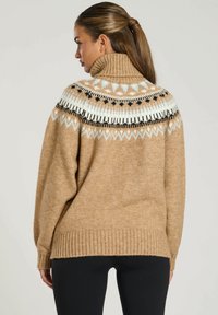 Beige stickad tröja med hög krage, med ett mönstrat ok i svart, vitt och ljusblått. Ribbad avslutning vid ärmar och nederkant.