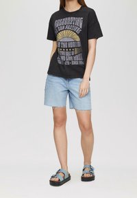 Osoba nosí černé tričko s grafikou, světle modré džíny s roztrhanými okraji a modré platformové sandály se dvěma přezkami.