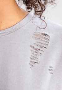 Gray sweatshirt featuring textured rips on one side, with a crew neckline and ribbed cuffs. Soft fabric with a smooth appearance.