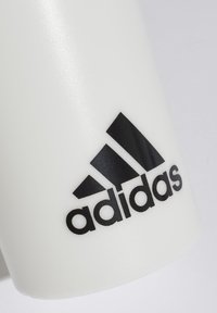 Objeto de plástico blanco con una textura mate que presenta un prominente logo negro de Adidas. El diseño incluye tres franjas diagonales por encima del logo.