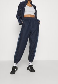 Kvinna som bär en marinblå träningsjacka och matchande byxor med elastiska muddar, grå crop top, vita strumpor och svarta Adidas-sneakers.