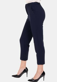 Donna che indossa pantaloni cropped slim-fit blu navy e décolleté nere in vernice, in piedi di lato su uno sfondo bianco.