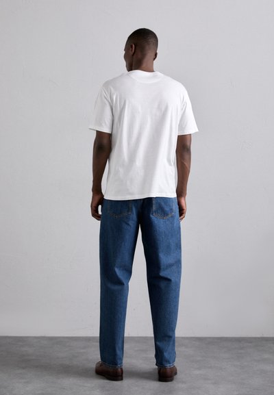 Camiseta de algodón blanca, con cuello redondo, mangas cortas, combinada con vaqueros de mezclilla azules con bolsillos y un corte relajado, llevada con zapatos marrones.