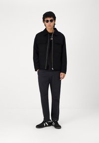 Μαύρο φερμουάρ μπουφάν με δύο μπροστινές τσέπες, συνδυασμένο με μαύρη μπλούζα, σκούρους γκρι παντελόνες και μαύρα αθλητικά παπούτσια με λευκές λεπτομέρειες.