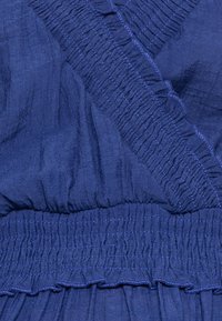 Tmavě modrá tkanina s texturovanými, nařasenými částmi. Vzory okrajů doplněné volánky a překřížený design u výstřihu.