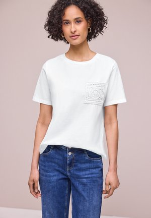 Kobieta o kręconych włosach, nosząca złote kolczyki-koła, białą koszulkę z krótkim rękawem i teksturowaną kieszonką oraz niebieskie dżinsy, stoi na jednolitym tle.