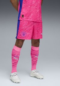 Różowy strój piłkarski z abstrakcyjnym wzorem, niebieskimi akcentami i emblematami. Zawiera pasujące różowe skarpety i białe korki z teksturowanym wzorem.