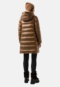 Frau, die einen glänzenden braunen Steppmantel mit Kapuze, schwarze Hosen, schwarze mit Fell gefütterte Stiefel und eine braune Strickmütze trägt, mit dem Rücken zur Kamera gewandt.