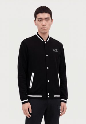 Hombre que lleva una chaqueta negra abotonada con rayas blancas en el cuello, los puños, los bolsillos y el logo EA7 Emporio Armani en el pecho.