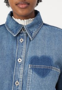 Denimskjorte med klassisk krage, sølvknapper og en hjerteformet, lys blå lapp på brystlommen. Teksturert stoff.
