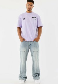 Lila oversized t-shirt met grafische accenten, gecombineerd met lichtblauwe baggy jeans met voorzakken en een vergeelte structuur. Witte sneakers.