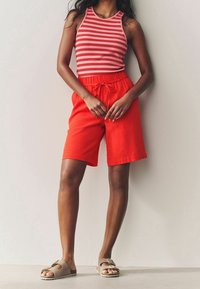 Rote Leinen-Shorts mit elastischem Bund und Kordelzug. Kombiniert mit einem ärmellosen Oberteil gestreift in Pink und Weiß sowie braunen Slippern.