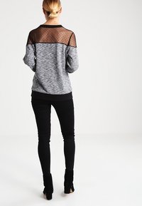 Grijze melange trui met een doorschijnende zwarte yoke met stippen. Gecombineerd met zwarte skinny jeans en enkellaarsjes, wat een gelaagde look laat zien.