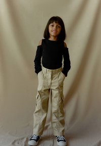 Zwarte lange mouw top met schouder uitsparingen, beige cargo broek met zijzakken, en zwarte sneakers met witte zolen. Neutrale achtergrond.