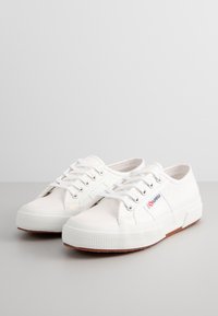 Sneaker bianche in tela con punta rotonda, suola in gomma testurizzata e dettagli argentati. Presentano un design con lacci e una patch con logo sul lato.