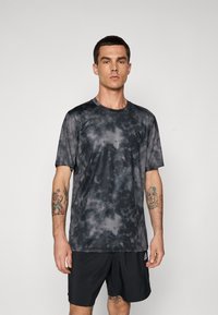 Mand iført en sort og grå mønstret atletisk T-shirt og sorte Adidas shorts, stående mod en ensfarvet lys grå baggrund.