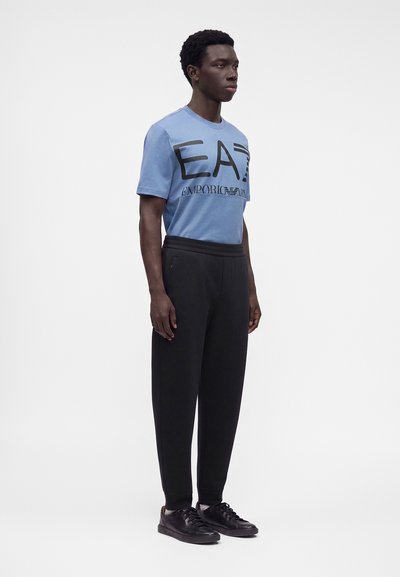 Άνδρας που φοράει μπλε μπλουζάκι Emporio Armani EA7, μαύρο παντελόνι και μαύρα αθλητικά παπούτσια σε λευκό φόντο.