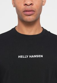 Close-up of person wearing black crewneck t-shirt with white "HELLY HANSEN" logo centered on the chest against a plain background.