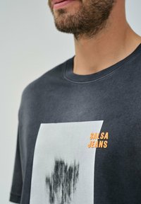 Camiseta de algodón gris oscuro con un gráfico blanco y un diseño negro, y texto naranja "SALSA JEANS" en el frente. Cuello redondo, mangas cortas.