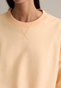 Sweat-shirt couleur pêche avec un col rond côtelé, une texture lisse, et un détail de couture en forme de 'X' sur l'avant. Design simple avec des accents minimalistes.