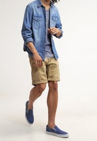 Chemise en denim avec poches à rabat, t-shirt gris, shorts beiges et chaussures à enfiler bleues. La tenue présente une coupe décontractée et un style casual.