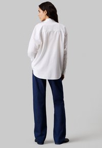 Camisa blanca de manga larga con un corte relajado y dobladillo curvado, combinada con pantalones de mezclilla de pierna ancha de color azul oscuro. Textura de tela suave.