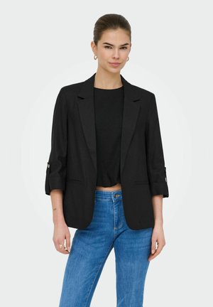 Jonge vrouw met haar naar achter gebonden, draagt gouden oorringen, een zwarte blazer met opgerolde mouwen, een zwarte top en blauwe jeans, staand tegen een witte achtergrond.