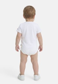 Νήπιο όρθιο με την πλάτη γυρισμένη, φορώντας ένα λευκό κορμάκι με κοντά μανίκια και λευκά παπούτσια σε ένα απλό λευκό φόντο.