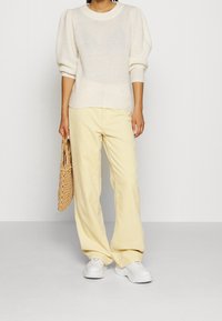 Sweter o kremowej teksturze z bufkami na rękawach, zestawiony z jasnosłomkowymi szerokimi spodniami z kordurowej tkaniny. Modelka trzyma plecioną torebkę w odcieniu beżu i nosi białe trampki.