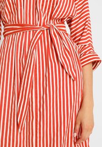 Rød- og hvidstribet kjole med bånd i taljen, korte ærmer og knapper foran. Fremstillet af let stof med en sprød tekstur.