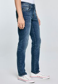 Jeans de denim ajustados en azul oscuro con un ligero desgastado, diseño de cinco bolsillos y costuras en contraste. Llevados con zapatillas blancas.