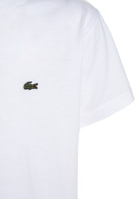 Biała bawełniana koszulka z krótkim rękawem, z małym wyszytym logo krokodyla w zielonym i czerwonym kolorze na lewej piersi.