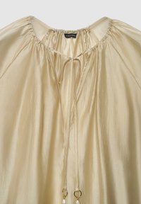 Cremefarbene, leichte Bluse mit rundem Halsausschnitt, gerafftem Stoff, Kordelzügen und zwei dekorativen Ringen an den Enden der Züge.