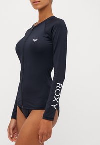 Rash guard a maniche lunghe di colore blu navy realizzato in materiale elastico, con zip frontale e il marchio bianco "ROXY" sulla manica sinistra.