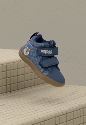 Baskets bleues pour enfants avec un personnage de Pat'Patrouille, deux sangles Velcro et une semelle marron clair, posées sur des marches carrelées sur fond uni.